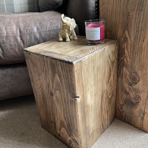 May include: A rustic wooden side table with a natural wood finish. The table is square and has a simple design. A gold elephant figurine and a candle with the label "Spiced Berries" are on top of the table.