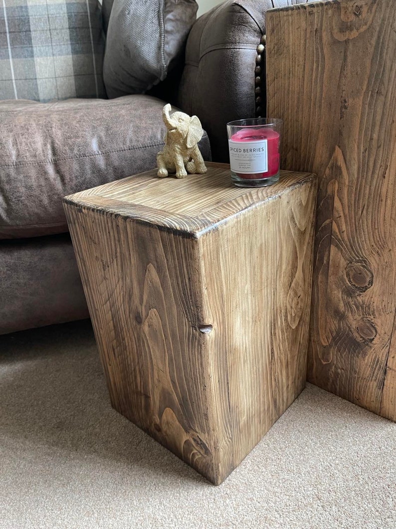 May include: A rustic wooden side table with a natural wood finish. The table is square and has a simple design. A gold elephant figurine and a candle with the label "Spiced Berries" are on top of the table.