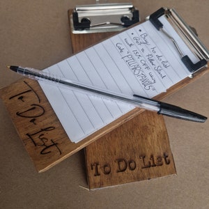 Op de afbeelding: Een houten clipboard met een to-do list erop geschreven. Het clipboard heeft een metalen clip en is gemaakt van donkerbruin hout. De tekst op het clipboard zegt "Buy to do list • Bet a Pillar Stand with 15% OFF using Cake "PILLARSTANDIS"" en "To Do List".