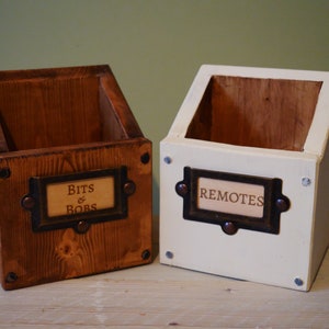Könnte beinhalten: Zwei Holzaufbewahrungsboxen, eine braun gebeizt und die andere weiß lackiert. Jede Box hat einen Etikettenhalter mit dem Text "BITS & BOBS" und "REMOTES". Die Boxen sind abgewinkelt und haben ein rustikales, handgefertigtes Aussehen.