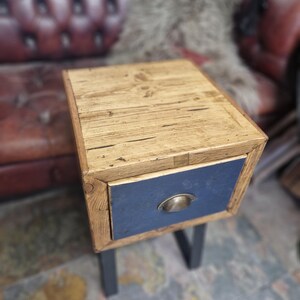 May include: A small, rustic wooden side table with a natural wood top and a navy blue drawer. The drawer has a brass-coloured handle. The table has dark grey legs.