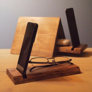 Op de afbeelding: Houten telefoon- en brillenstandaards. De standaards zijn gemaakt van donkerbruin hout en hebben een eenvoudig, minimalistisch ontwerp. Eén standaard houdt een bril vast, terwijl de andere een telefoon vasthoudt. De achtergrond is een lichtblauwe muur.