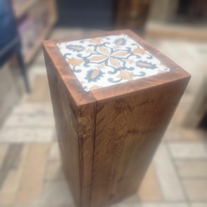 Op de afbeelding: Een houten kruk met een vierkante bovenkant met een decoratieve tegel met een bloemmotief in de kleuren blauw, wit en goud.
