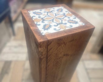 May include: A wooden stool with a square top featuring a decorative tile with a floral pattern in shades of blue, white, and gold.