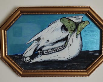 Könnte beinhalten: Ein achteckiges gerahmtes Kunstwerk mit einem weißen Pferdeschädel mit detaillierten Zähnen und einem grünen Schmetterling. Der Hintergrund ist ein Farbverlauf aus Blau und Schwarz. Der Rahmen ist goldfarben.