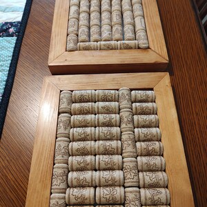 Può includere: Due cornici rettangolari in legno contenenti composizioni di tappi di sughero naturali. Ogni tappo presenta una grafica stampata di viti e grappoli d'uva. Le cornici sono realizzate in legno chiaro con una finitura liscia e lucida.