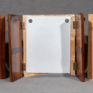 May include: A wooden display case with three hinged panels. The panels are made of different types of wood, including a dark reddish-brown, a lighter brown, and a natural wood color. The case is open, revealing a white interior.