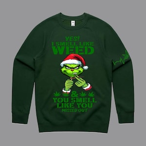 May include: Dark green sweatshirt with a graphic of a green character wearing a Santa hat, holding a lit cigarette. Text reads "YES! I SMELL LIKE WEED & YOU SMELL LIKE YOU MISSED OUT."
