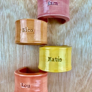 May include: Four ceramic napkin rings in different colors, with the names "Sam", "Nico", "Katie", and "Lou" written on them.