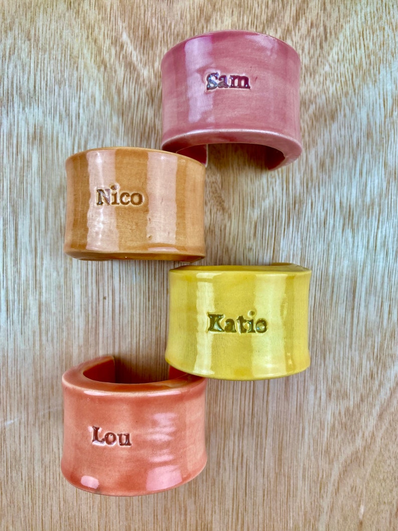 May include: Four ceramic napkin rings in different colors, with the names "Sam", "Nico", "Katie", and "Lou" written on them.