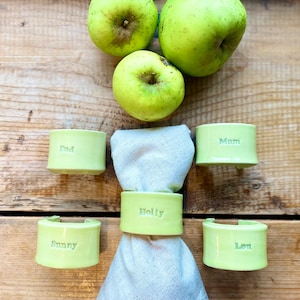 May include: Five green ceramic napkin rings with names inscribed on each. The names are "Dad", "Mum", "Holly", "Sunny", and "Lou". Three green apples are in the background.