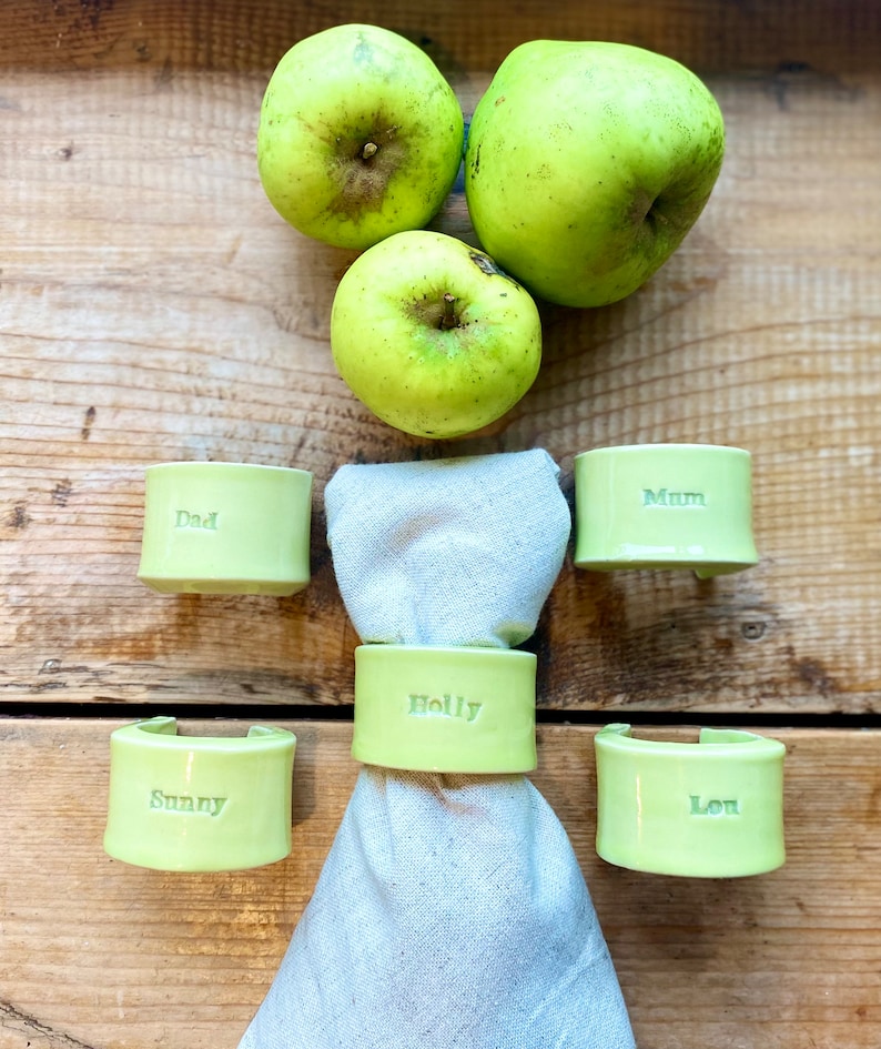 May include: Five green ceramic napkin rings with names inscribed on each. The names are "Dad", "Mum", "Holly", "Sunny", and "Lou". Three green apples are in the background.