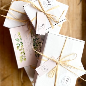 May include: Three white gift boxes tied with natural twine and decorated with a small, round, white sticker with the text "NY Ceramics" and a black circle with the letter "N" inside. The boxes are stacked on top of each other. A pink card with green foliage and the text "Thank you for your order" is visible in the front.
