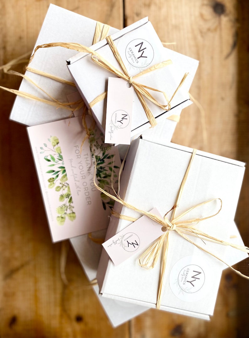 May include: Three white gift boxes tied with natural twine and decorated with a small, round, white sticker with the text "NY Ceramics" and a black circle with the letter "N" inside. The boxes are stacked on top of each other. A pink card with green foliage and the text "Thank you for your order" is visible in the front.