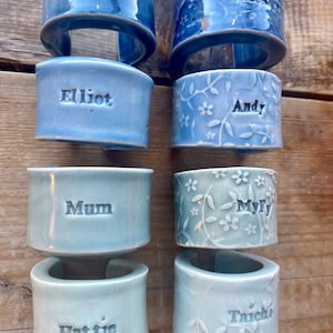 May include: Eight ceramic napkin rings with different names and designs. The rings are in shades of blue and green. The names on the rings are Dad, Mark, Elliot, Andy, Mum, Myf, Hattie, and Taichi.