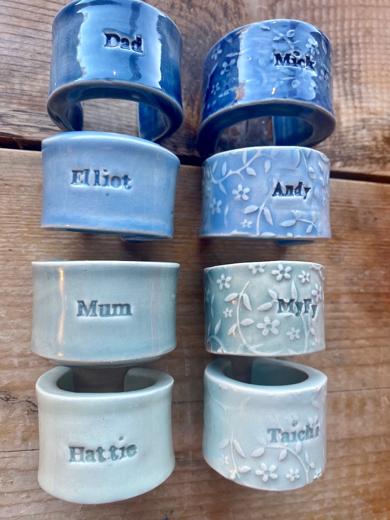 May include: Eight ceramic napkin rings with different names and designs. The rings are in shades of blue and green. The names on the rings are Dad, Mark, Elliot, Andy, Mum, Myf, Hattie, and Taichi.