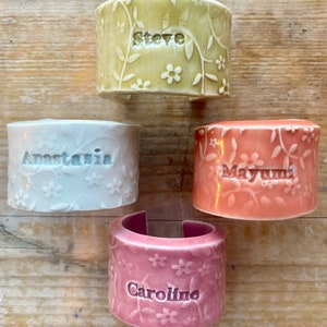 May include: Four ceramic napkin rings with floral designs and personalized names. The rings are in shades of green, white, pink, and orange.