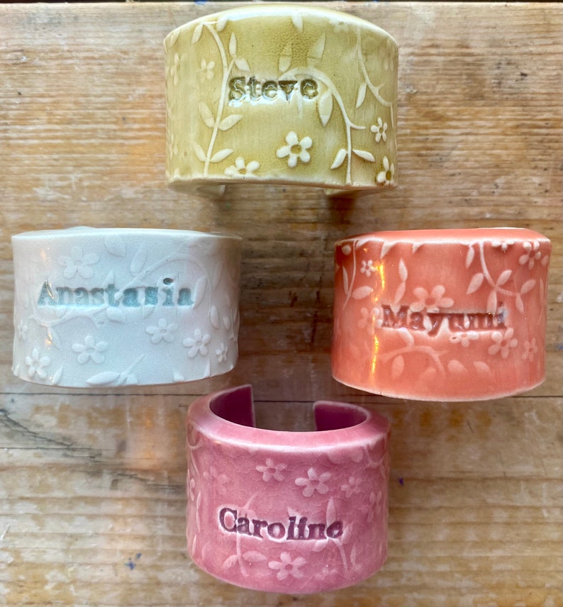 May include: Four ceramic napkin rings with floral designs and personalized names. The rings are in shades of green, white, pink, and orange.