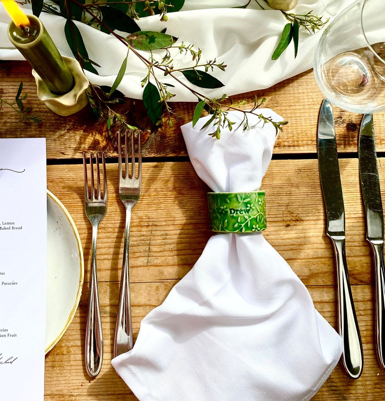 May include: A white napkin with a green ceramic napkin ring that says "Drew" on it. The napkin ring has a floral design. The napkin is on a wooden table with a fork and knife beside it.