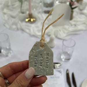 Könnte beinhalten: Hellblaue Keramik-Anhänger mit dem geprägten Schriftzug "Mother of the Bride". Der Anhänger hat ein Blumenmuster und ist mit einer Bastkordel gebunden. Der Anhänger wird in einer Hand gehalten, mit einer Hochzeitstafel im Hintergrund.