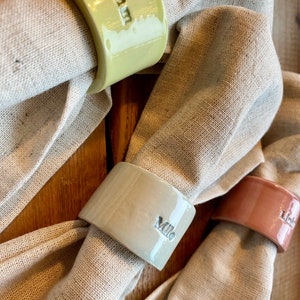 May include: Three ceramic napkin rings with personalized names. The rings are in shades of green, beige, and pink. The names are "Olivia", "Milo", and "Lily".