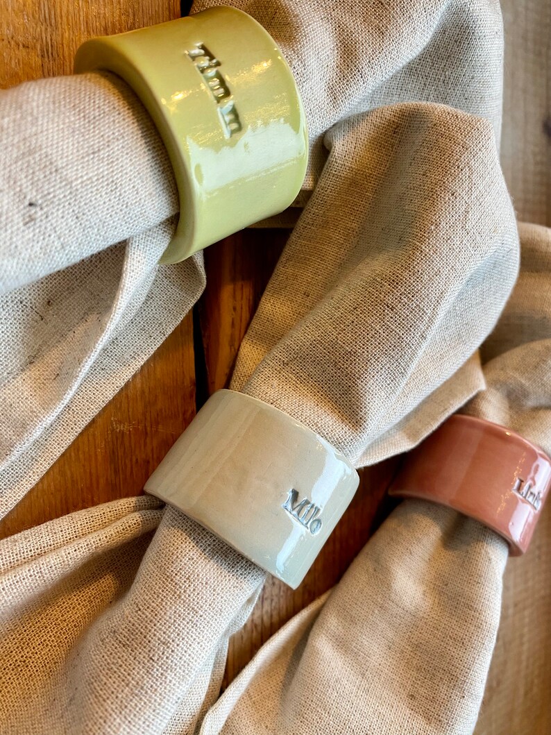 May include: Three ceramic napkin rings with personalized names. The rings are in shades of green, beige, and pink. The names are "Olivia", "Milo", and "Lily".