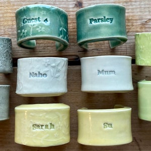 May include: Ten ceramic napkin rings with different names and designs. The rings are in various shades of green, white, and yellow. The names on the rings include "Guest 4", "Parsley", "Ilias", "Naho", "Mum", "Melie", "Robertson", "Sarah", "Su", and "Namaste".