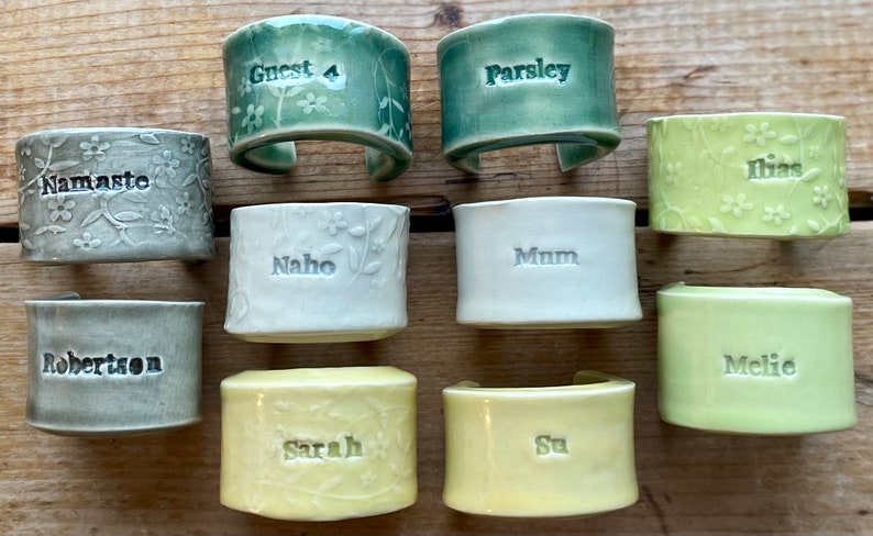 May include: Ten ceramic napkin rings with different names and designs. The rings are in various shades of green, white, and yellow. The names on the rings include "Guest 4", "Parsley", "Ilias", "Naho", "Mum", "Melie", "Robertson", "Sarah", "Su", and "Namaste".