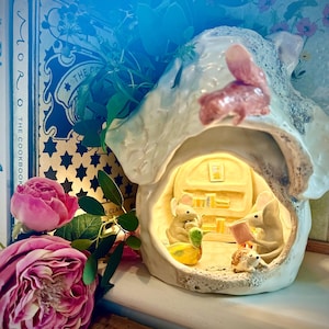 May include: A ceramic house-shaped night light featuring a squirrel and a warm interior scene. Two mice read books, with a duck and a hedgehog. Pink roses and a cookbook are in the background, creating a charming display.