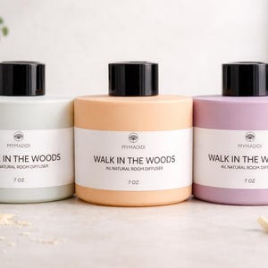Könnte beinhalten: Drei zylindrische Raumduftdiffusoren in Pastellfarben: Hellgrün, Pfirsich und Lavendel. Jeder hat einen schwarzen Deckel und ein weißes Etikett mit dem Text "WALK IN THE WOODS" und "7 OZ". Ein Produkt für Raumduft.