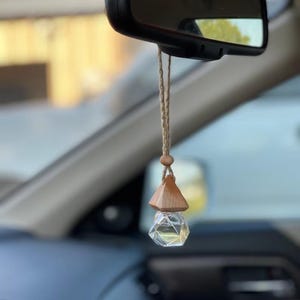 May include: A car air freshener hanging from a rearview mirror, featuring a clear, multi-faceted glass bottle with a geometric shape, topped with a light-colored wooden cap and suspended by a thin, braided tan cord with a small wooden bead.