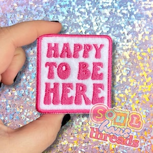 May include: A square, white patch with a pink border and the words "HAPPY TO BE HERE" embroidered in pink, stylized letters. The patch is held up against a sparkly, iridescent background. The patch is a product of Soul Haus Threads.