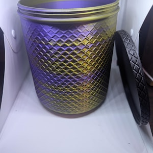 Op de afbeelding: Een cilindrische container met een deksel. De container heeft een verloop van paars en goud, met een ruitpatroon. Het deksel is zwart en heeft een bijpassend ruitpatroon. De container staat op een wit oppervlak.