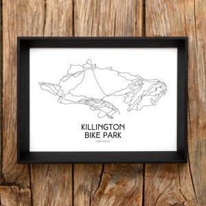 May include: Black framed print featuring a minimalist line drawing of a mountain bike park map. The text "KILLINGTON BIKE PARK Vermont" is printed in black below the map. The print is set against a wooden background.