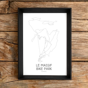 May include: A framed black and white line art print of a mountain bike park map. The print features the text "LE MASSIF BIKE PARK Quebec" in a minimalist style. The frame is black, and the background is a wooden surface.
