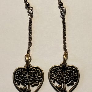 Puede incluir: Un par de pendientes colgantes con una cadena de metal en tono bronce unida a un colgante en forma de corazón. El colgante presenta un diseño recortado de un árbol con un pequeño corazón en la base. Los pendientes incluyen ganchos metálicos.