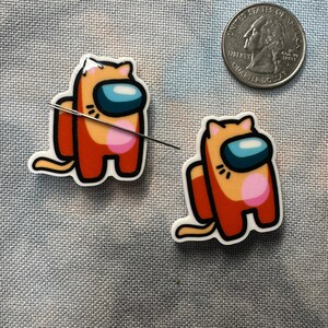May include: Two orange and pink cat-themed Among Us character stickers. The stickers are on a gray fabric background.