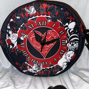 May include: A black and red round crossbody bag with a white clock face and a red heart design. The bag features characters from Alice in Wonderland, including the Queen of Hearts, the Mad Hatter, and Alice. The bag has an adjustable strap.