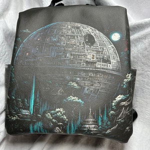 May include: A black leather backpack with a detailed illustration of the Death Star from Star Wars. The illustration is in shades of gray and blue, with a white moon in the background.