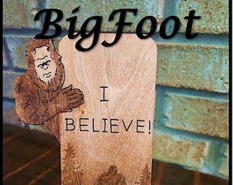 May include: Wooden cell phone stand with a Bigfoot design. The stand features a Bigfoot character holding a sign that says "I Believe!" and a silhouette of Bigfoot in the woods.