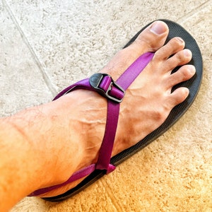 May include: A black sandal with a purple strap and a black buckle. The sandal is on a foot. The foot is on a light-colored tile floor.