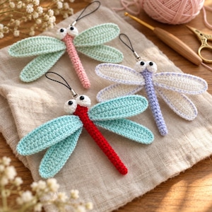 May include: Three handmade crochet dragonfly ornaments with black hanging loops. One has a pink body and mint wings, one is lavender with white wings, and one features a bright red body with light blue wings. Each has two white round eyes.