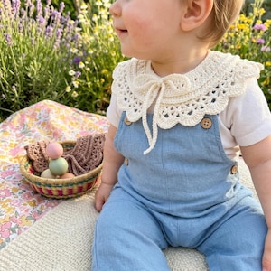 May include: A baby wearing a light blue denim jumpsuit with wooden buttons and a cream-colored, crocheted collar. A small basket with wooden toys sits on a floral blanket. The scene is set outdoors with flowers in the background.