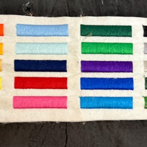 Może przedstawiać: Próbka tkaniny z szeregiem kolorowych wstążek. Wstążki ułożone są w rzędach i kolumnach, prezentując różnorodne odcienie, w tym pomarańczowy, żółty, niebieski, zielony i różowy. Próbka znajduje się na neutralnym tle.