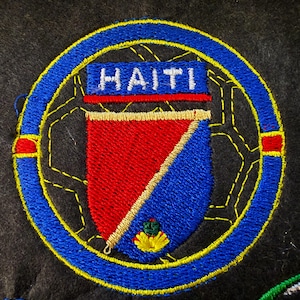 May include: Embroidered patch featuring the word "HAITI" in white on a blue rectangle. The patch has a shield design with red and blue sections, surrounded by a blue circle and yellow stitching. The background is dark.