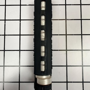 May include: A black cylindrical object with a textured surface and three rectangular openings revealing white thread spools. The object has a silver top and bottom, and is positioned vertically against a grid background.