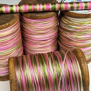 May include: Four wooden spools wrapped with variegated thread in shades of pink, olive green, and white. The thread features a shiny, metallic-like finish and is wound tightly around each spool. One spool is positioned horizontally in front.