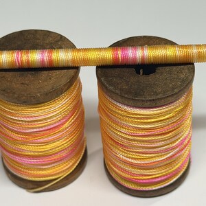 May include: Two vintage wooden spools wrapped with variegated thread in shades of yellow, orange, and pink. A single wooden dowel is positioned horizontally across the tops of both spools, also wrapped tightly with the same multicolored thread.