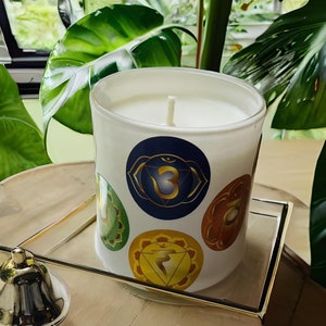 May include: White candle in a glass jar with a white lid. The jar has colourful chakra symbols printed on it. The candle is on a wooden surface with a gold-coloured tray.