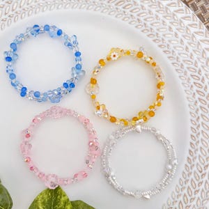 May include: Four beaded bracelets in shades of blue, yellow, pink, and clear. The bracelets feature various bead shapes, including flowers and hearts. The bracelets are arranged on a white plate with a decorative edge.
